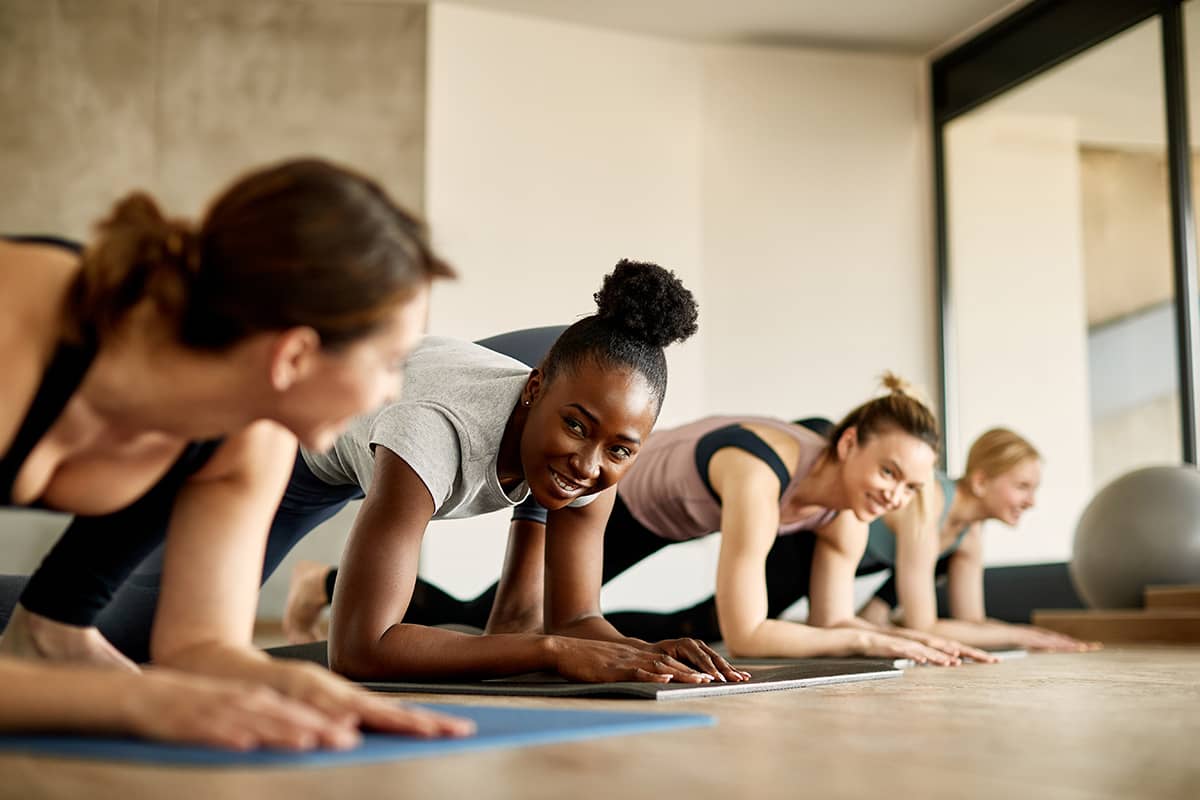 group-class-01 Women on yoga mats hold planks and smile at each other during a group fitness class